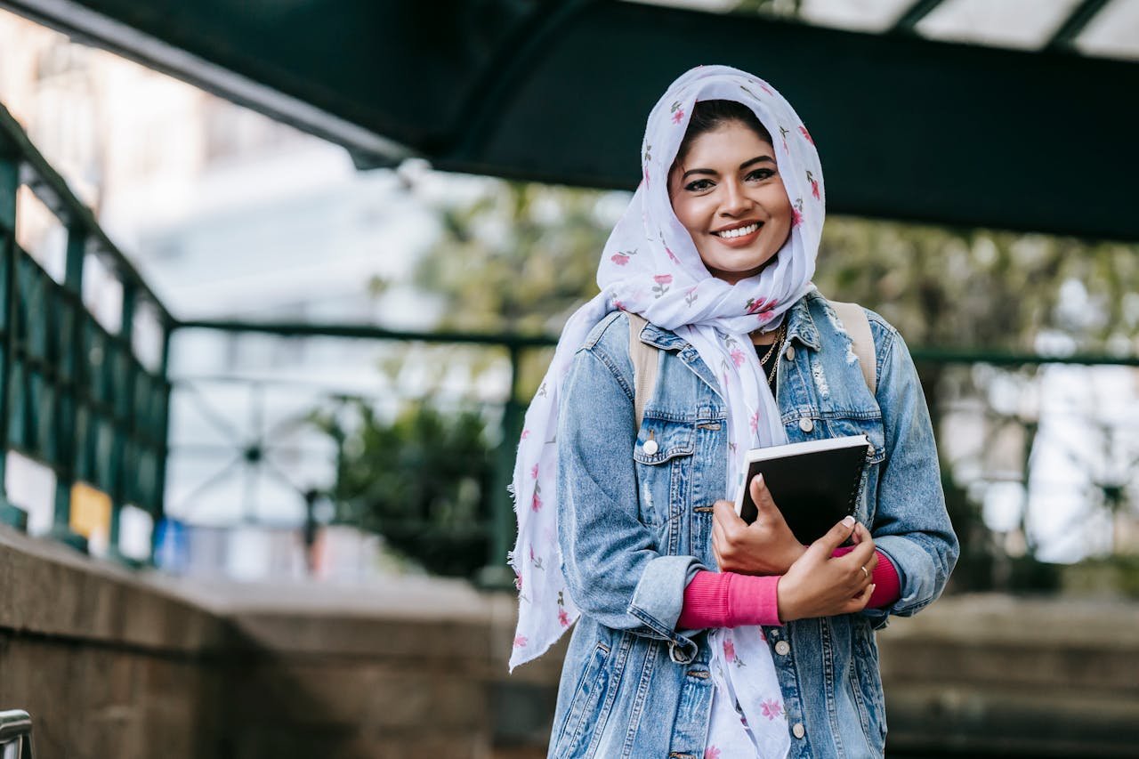 Content ethnic female in headscarf and denim coat with notebook looking at camera with toothy smile