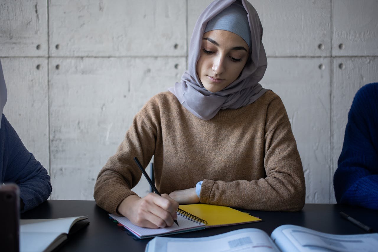 Focused Muslim woman in hijab writing in notebook during a study session indoors.