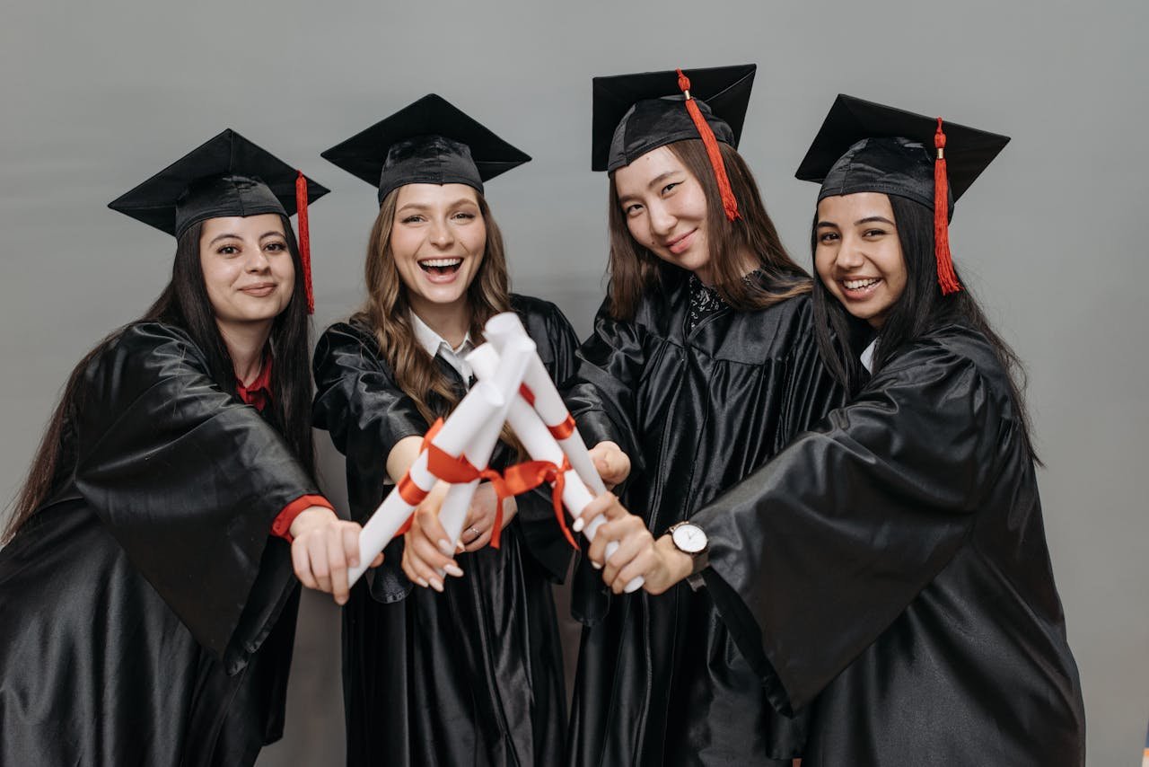 Offerings A joyful group of multiracial women in graduation gowns holding diplomas.