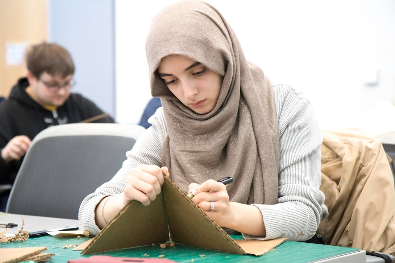 Young Muslim woman wearing hijab working on a cardboard craft project in classroom setting.